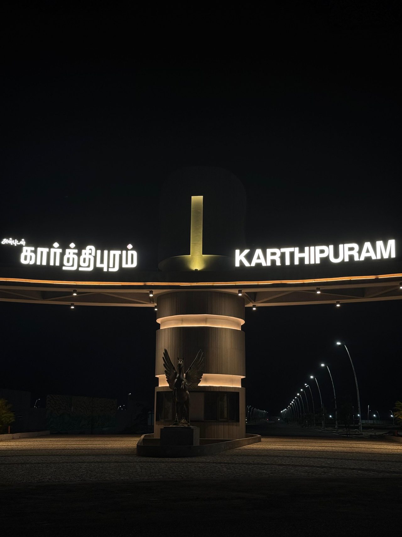 Karthipuram township entrance gateway lit at night, with the avenue of street poles beyond